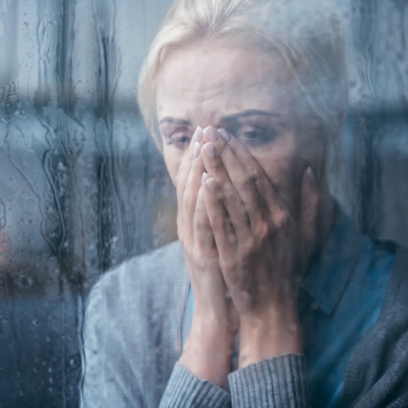 sad adult woman crying and covering face with hands at home through window with raindrops